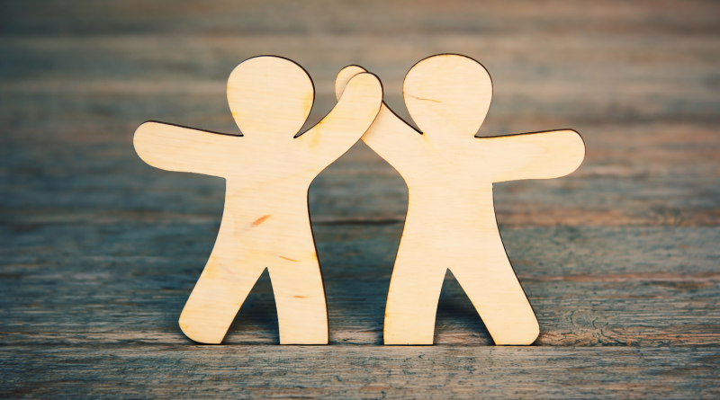 Deux silhouettes humaines en bois se touchant la main dans un geste de solidarité, posées sur une surface en bois.