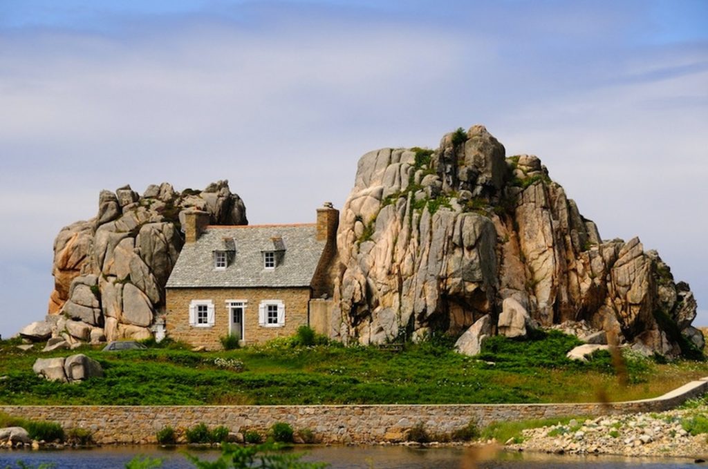 Maison en pierre isolée nichée entre deux immenses formations rocheuses granitiques, au cœur d’un paysage côtier verdoyant sous un ciel clair.