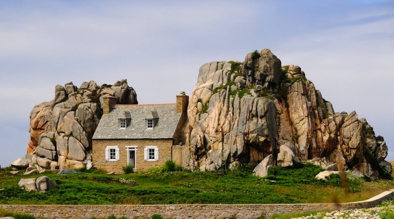 Maison en pierre isolée nichée entre deux immenses formations rocheuses granitiques, au cœur d’un paysage côtier verdoyant sous un ciel clair.