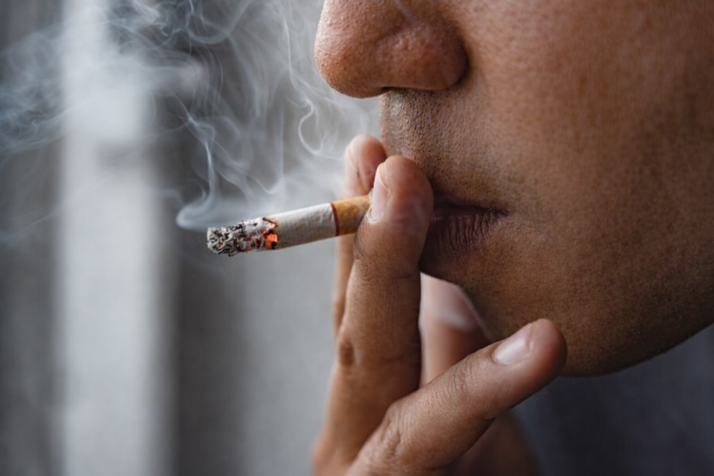 Gros plan d’un visage en train de fumer une cigarette, avec la braise allumée et la fumée visible près des lèvres.