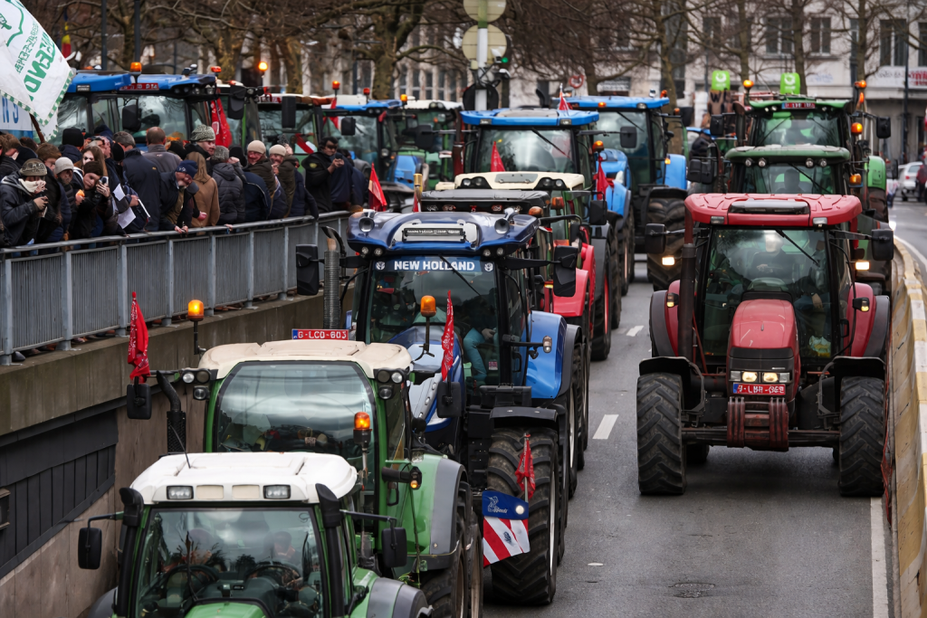 Convoi de tracteurs bloquant une avenue urbaine, foule de manifestants sur le côté, gyrophares allumés.