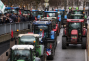 Convoi de tracteurs bloquant une avenue urbaine, foule de manifestants sur le côté, gyrophares allumés.