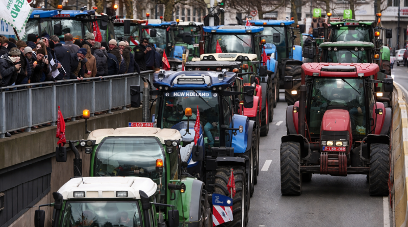 Convoi de tracteurs bloquant une avenue urbaine, foule de manifestants sur le côté, gyrophares allumés.