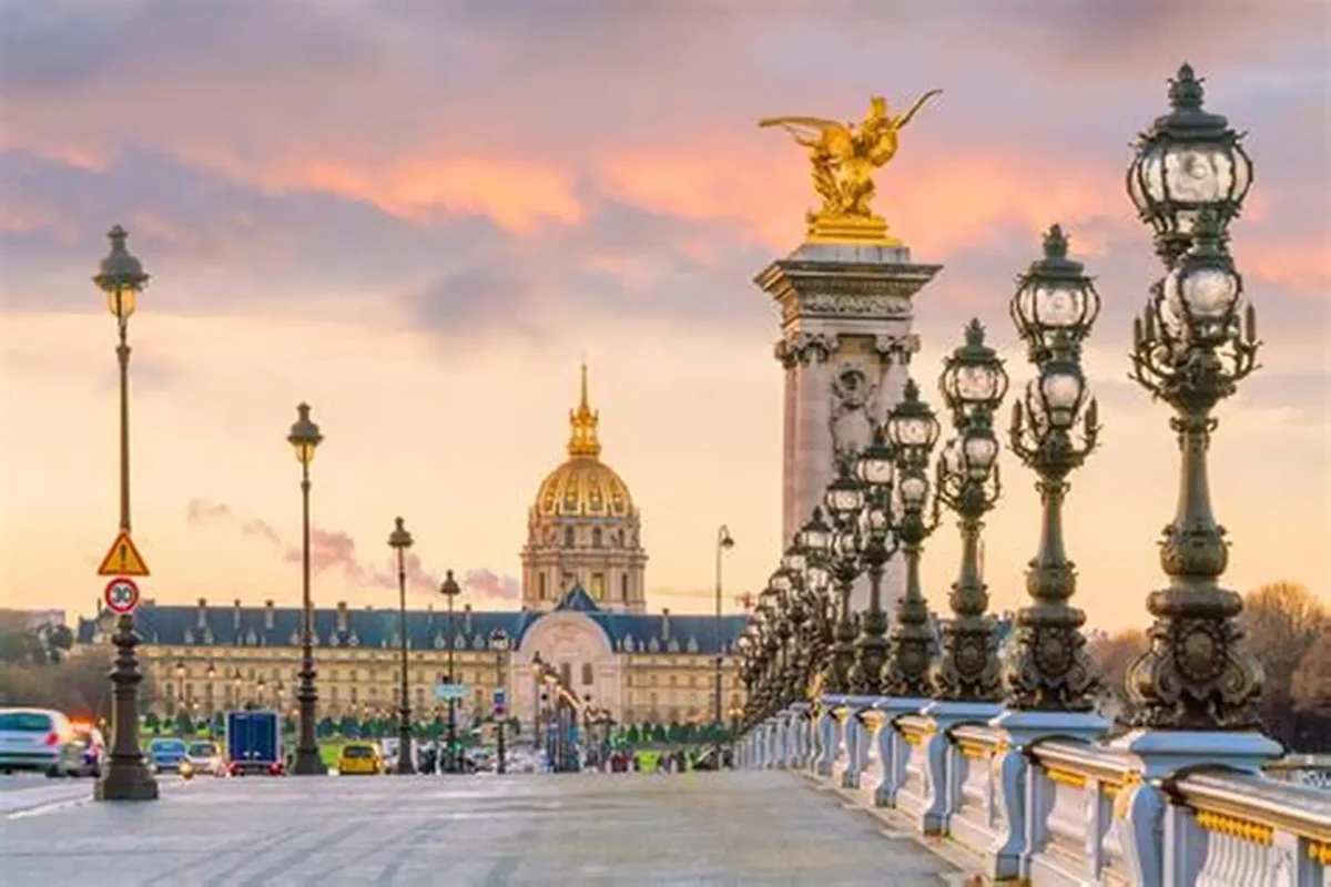 Vue panoramique du pont Alexandre III à Paris au coucher du soleil, avec ses lampadaires en fonte richement décorés alignés le long du pont, une statue dorée ailée au sommet d’un pilier monumental, quelques véhicules circulant au loin et le dôme doré des Invalides en arrière-plan sous un ciel aux teintes roses et orangées.