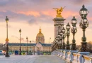Vue panoramique du pont Alexandre III à Paris au coucher du soleil, avec ses lampadaires en fonte richement décorés alignés le long du pont, une statue dorée ailée au sommet d’un pilier monumental, quelques véhicules circulant au loin et le dôme doré des Invalides en arrière-plan sous un ciel aux teintes roses et orangées.