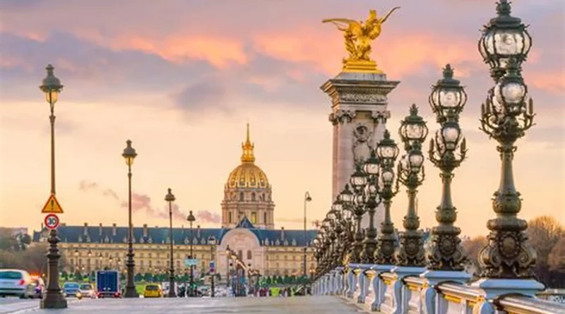 Vue panoramique du pont Alexandre III à Paris au coucher du soleil, avec ses lampadaires en fonte richement décorés alignés le long du pont, une statue dorée ailée au sommet d’un pilier monumental, quelques véhicules circulant au loin et le dôme doré des Invalides en arrière-plan sous un ciel aux teintes roses et orangées.