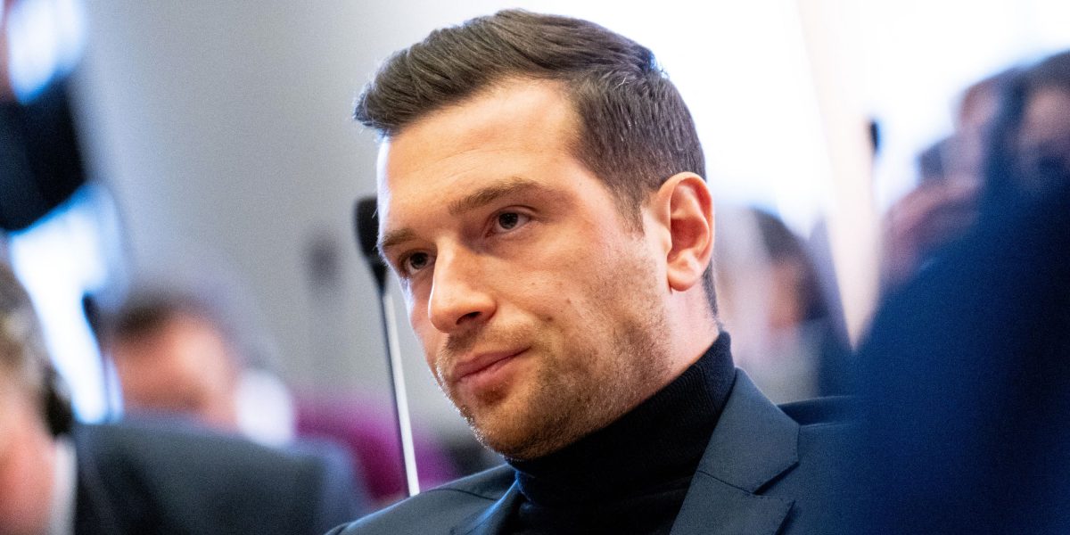 Homme assis dans un environnement institutionnel, vêtu d’un costume sombre, regard concentré, entouré de personnes floues à l’arrière-plan, illustrant une scène de débat ou de réunion publique