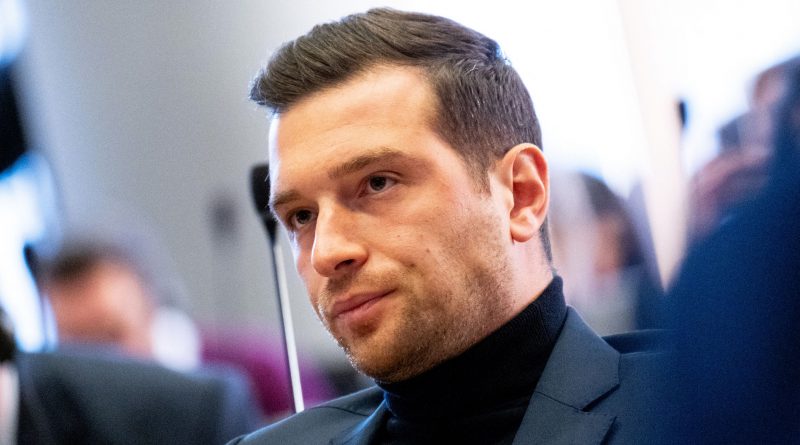 Homme assis dans un environnement institutionnel, vêtu d’un costume sombre, regard concentré, entouré de personnes floues à l’arrière-plan, illustrant une scène de débat ou de réunion publique