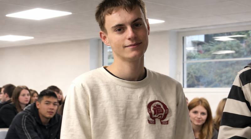 Jeune homme debout dans une salle de classe, regardant l’objectif, avec des élèves assis en arrière-plan sous un éclairage intérieur.