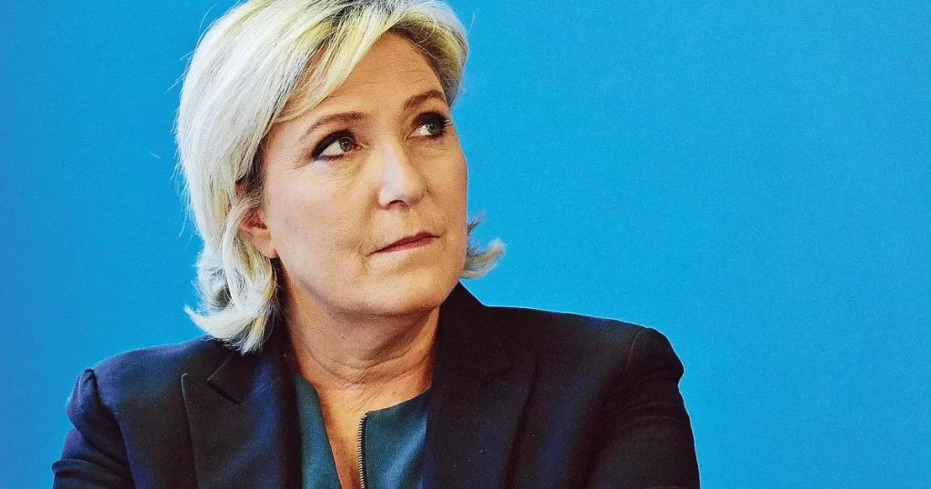 Femme politique blonde au regard sérieux, photographiée de profil devant un fond bleu uni, posture concentrée et ferme