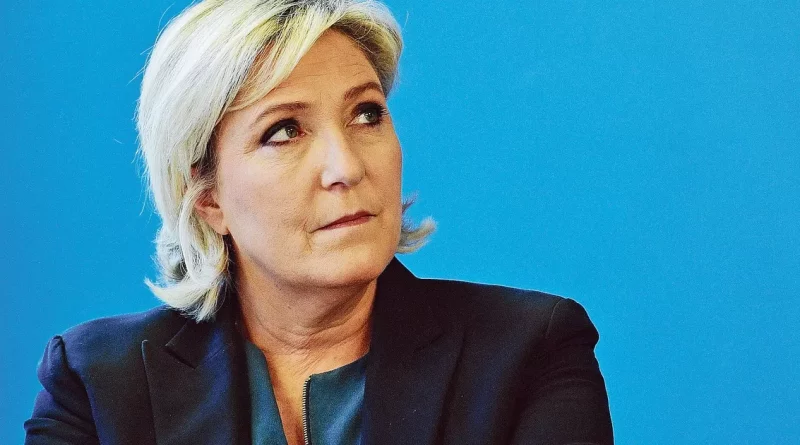 Femme politique blonde au regard sérieux, photographiée de profil devant un fond bleu uni, posture concentrée et ferme