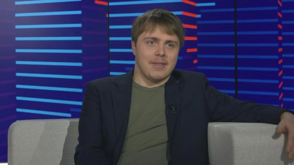 Jeune homme assis sur un canapé lors d’une émission en plateau télévisé, regard tourné vers l’interlocuteur, expression calme et attentive, fond composé de lumières bleues et rouges floues