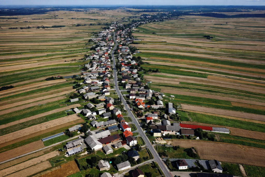 Vue aérienne d’un village rural très allongé, organisé le long d’une route unique traversant des champs agricoles aux formes régulières et colorées.