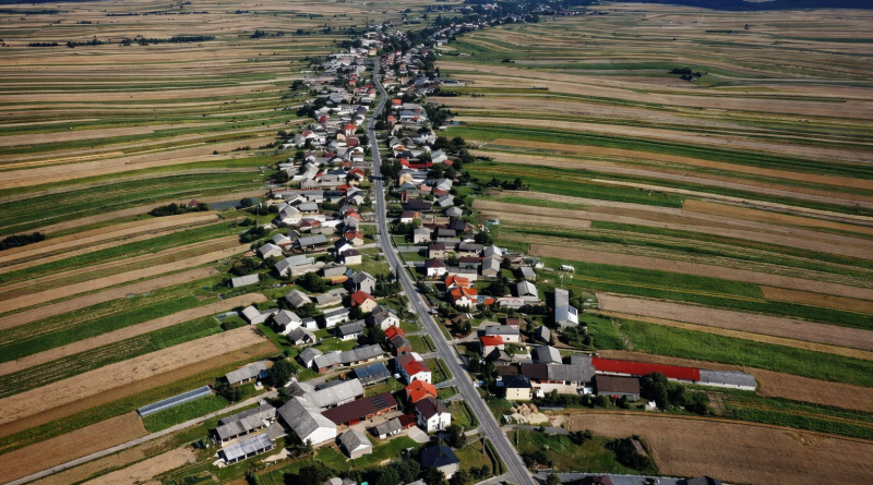 Vue aérienne d’un village rural très allongé, organisé le long d’une route unique traversant des champs agricoles aux formes régulières et colorées.