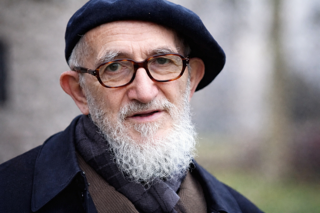 Portrait d’un homme âgé portant un béret sombre et des lunettes rondes, barbe blanche fournie, regard attentif, arrière-plan extérieur flou.