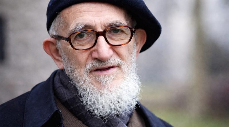 Portrait d’un homme âgé portant un béret sombre et des lunettes rondes, barbe blanche fournie, regard attentif, arrière-plan extérieur flou.