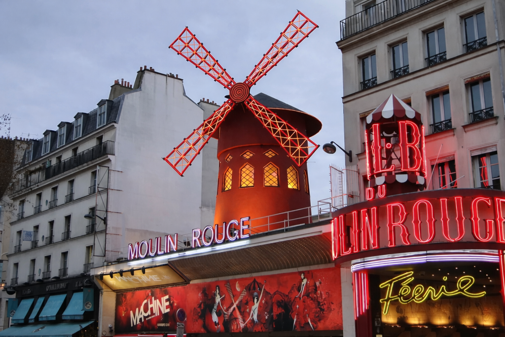 Façade illuminée d’un cabaret parisien emblématique avec un grand moulin rouge lumineux, enseignes néon allumées et immeubles urbains en arrière-plan, en début de soirée.