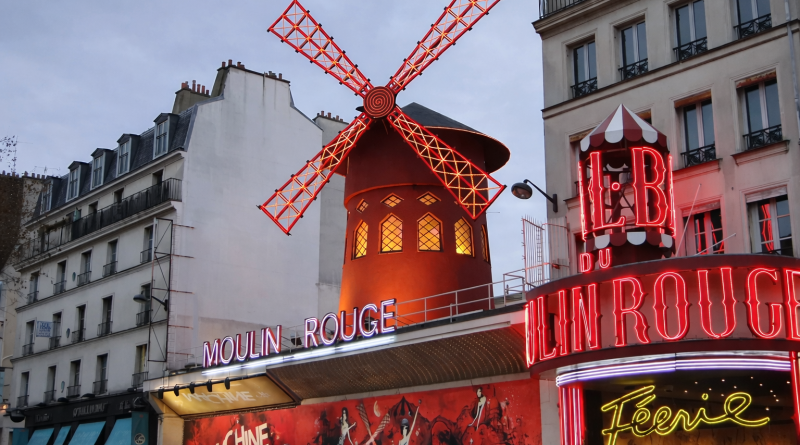 Façade illuminée d’un cabaret parisien emblématique avec un grand moulin rouge lumineux, enseignes néon allumées et immeubles urbains en arrière-plan, en début de soirée.