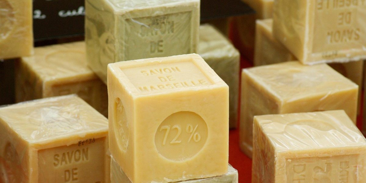 Cube de savon artisanal de couleur beige, empilé avec d’autres savons solides, présentant un marquage en relief indiquant une forte teneur en huiles végétales.