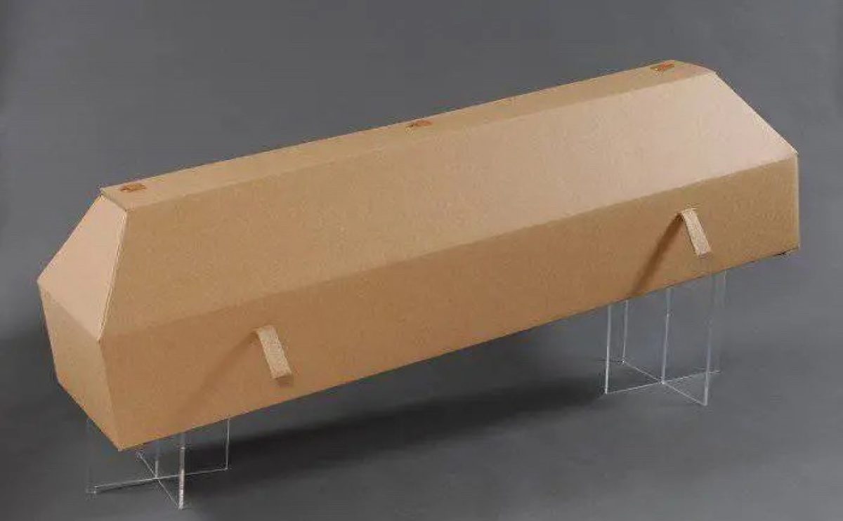 Cercueil en carton de couleur kraft, forme rectangulaire, présenté sur supports transparents, fond gris neutre.