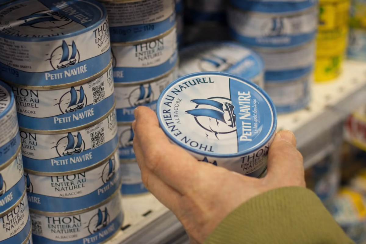 Main tenant une conserve de thon dans un rayon de supermarché, illustrant les inquiétudes liées à la présence de substances toxiques dans l’alimentation courante.