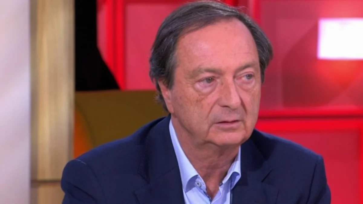 Homme d’âge mûr assis sur un plateau de télévision lors d’une interview, portant une veste sombre et une chemise claire, avec un décor de studio aux lumières rouges en arrière-plan.