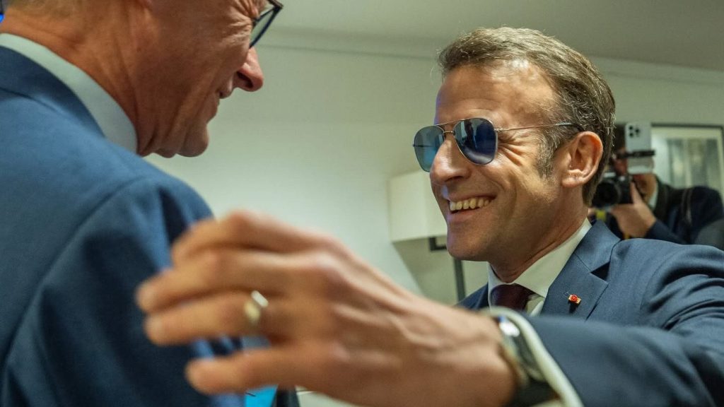 Un homme politique en costume sombre sourit et serre la main d’un autre responsable lors d’un échange en coulisses, sous l’œil des caméras, dans une atmosphère institutionnelle.