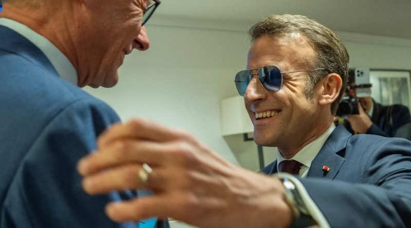 Un homme politique en costume sombre sourit et serre la main d’un autre responsable lors d’un échange en coulisses, sous l’œil des caméras, dans une atmosphère institutionnelle.