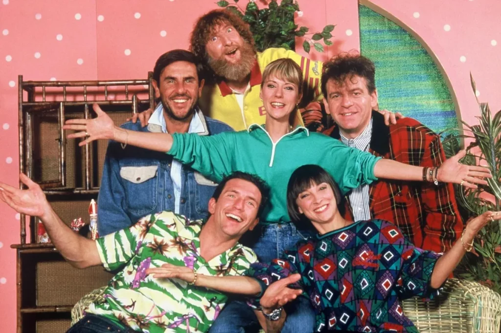 Groupe d’animateurs souriants posant ensemble sur un plateau de télévision coloré, décor rétro aux tons roses et verts, ambiance joyeuse et conviviale des années 1990.