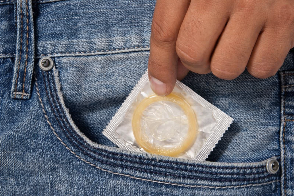 Main masculine tenant un préservatif emballé, partiellement sorti de la poche d’un jean, symbole de protection et de prévention.