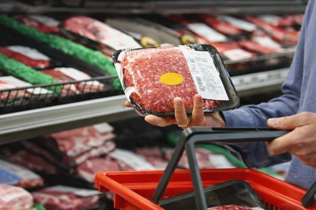 Barquette de viande hachée emballée tenue par un consommateur dans un rayon réfrigéré de supermarché, avec une étiquette visible sur l’emballage.