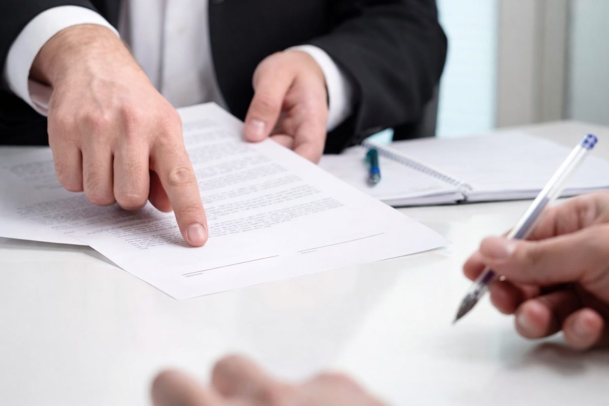 Main d’un homme en costume pointant une ligne sur un contrat de location posé sur une table, tandis qu’une autre personne tient un stylo prête à signer le document lié à un engagement de caution pour une location de logement.