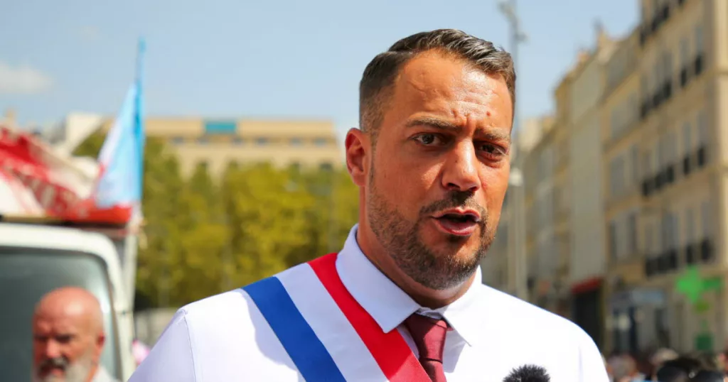 homme politique français en costume s’exprimant devant des micros avec une foule et des caméras en arrière-plan dans une ambiance tendue