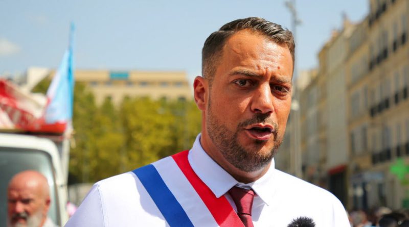 homme politique français en costume s’exprimant devant des micros avec une foule et des caméras en arrière-plan dans une ambiance tendue