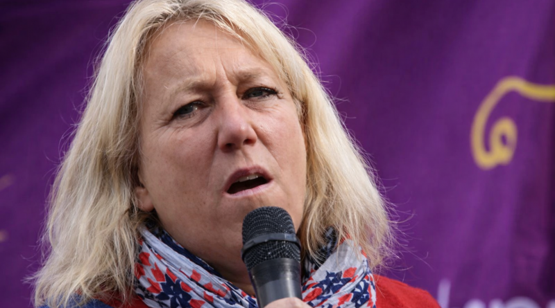 Femme blonde s’exprimant au micro lors d’un rassemblement, visage grave, fond violet flouté.