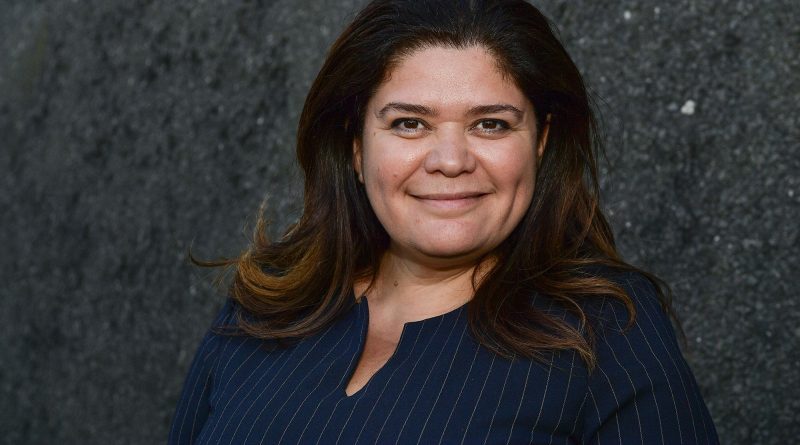 Portrait d’une femme brune souriante, regard face caméra, devant un mur sombre texturé.