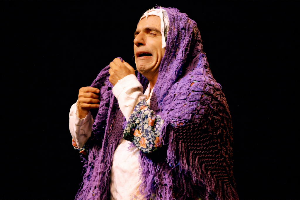 Personnage de scène enveloppé dans un châle violet, exprimant une forte émotion sous un éclairage théâtral sur fond sombre.