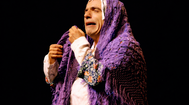 Personnage de scène enveloppé dans un châle violet, exprimant une forte émotion sous un éclairage théâtral sur fond sombre.