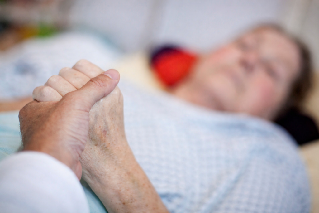 Main d’une personne tenant doucement celle d’une personne âgée allongée dans un lit médicalisé, dans une atmosphère calme et émotive.