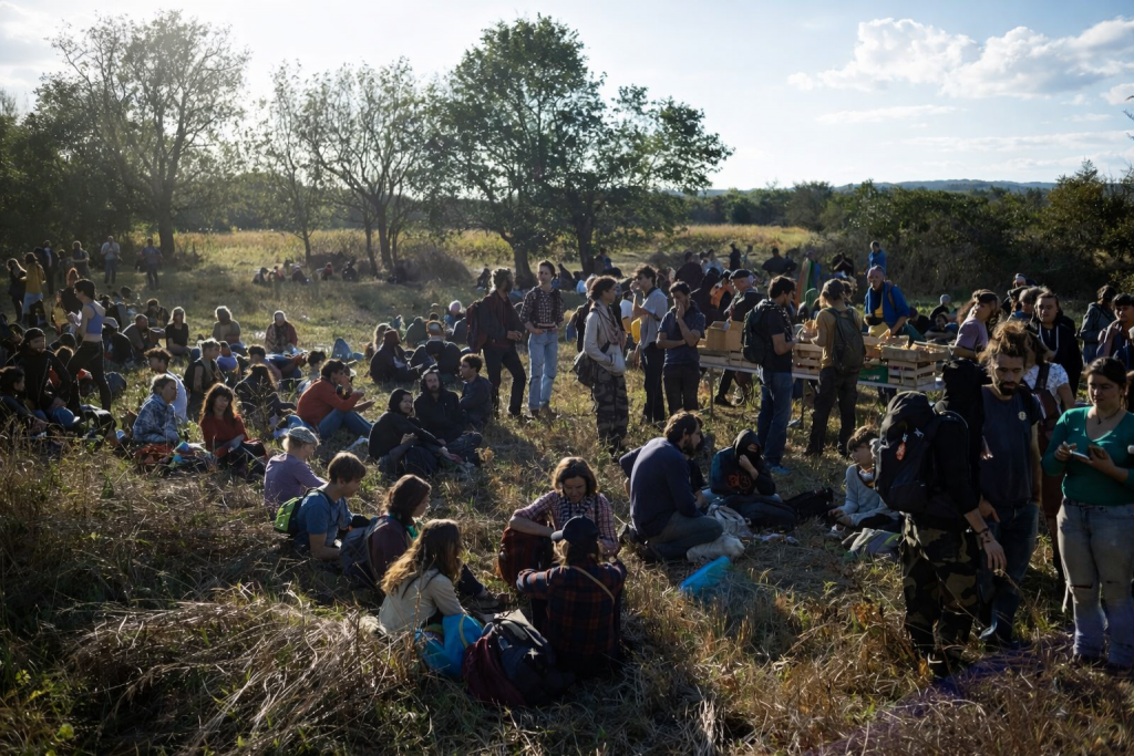 Groupe de personnes réunies dans un champ naturel, assises et debout en petits groupes, lors d’un rassemblement en plein air sous une lumière de fin de journée.