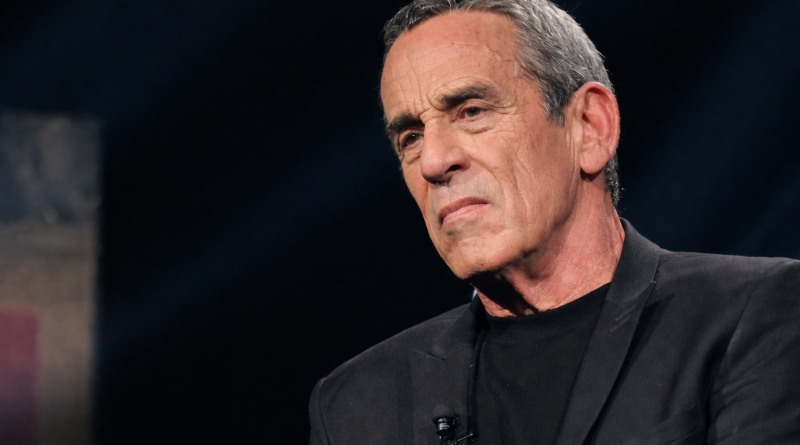 portrait serré d’un homme aux cheveux gris vêtu de noir, éclairage sombre et expression grave