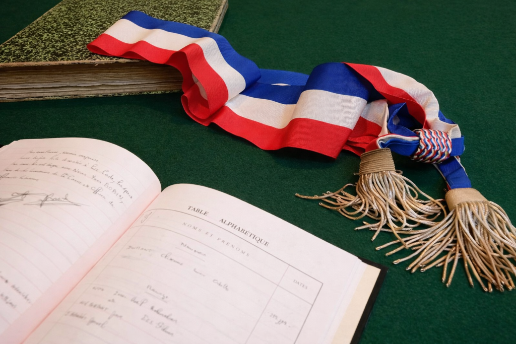 Écharpe tricolore posée sur une table verte à côté d’un registre ouvert et d’un ancien livre, symbole des institutions et de la vie démocratique locale.