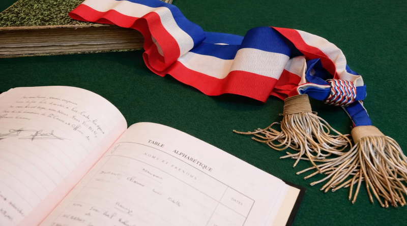 Écharpe tricolore posée sur une table verte à côté d’un registre ouvert et d’un ancien livre, symbole des institutions et de la vie démocratique locale.
