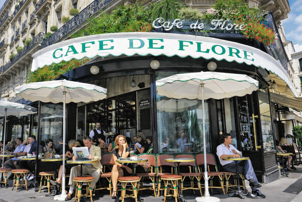 terrasse de café parisien animée avec tables rondes, parasols clairs et immeubles haussmanniens en arrière-plan