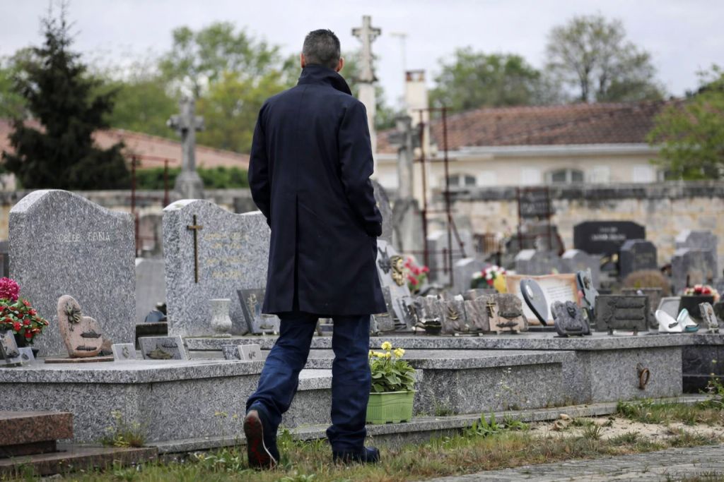 Homme de dos marchant dans un cimetière, entouré de tombes en pierre et de fleurs, sous un ciel gris et une atmosphère silencieuse.