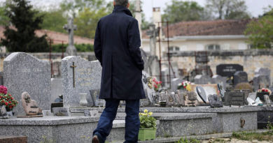 Homme de dos marchant dans un cimetière, entouré de tombes en pierre et de fleurs, sous un ciel gris et une atmosphère silencieuse.