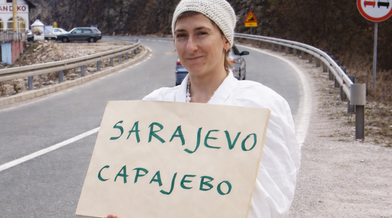femme vêtue de blanc tenant une pancarte manuscrite au bord d’une route de montagne