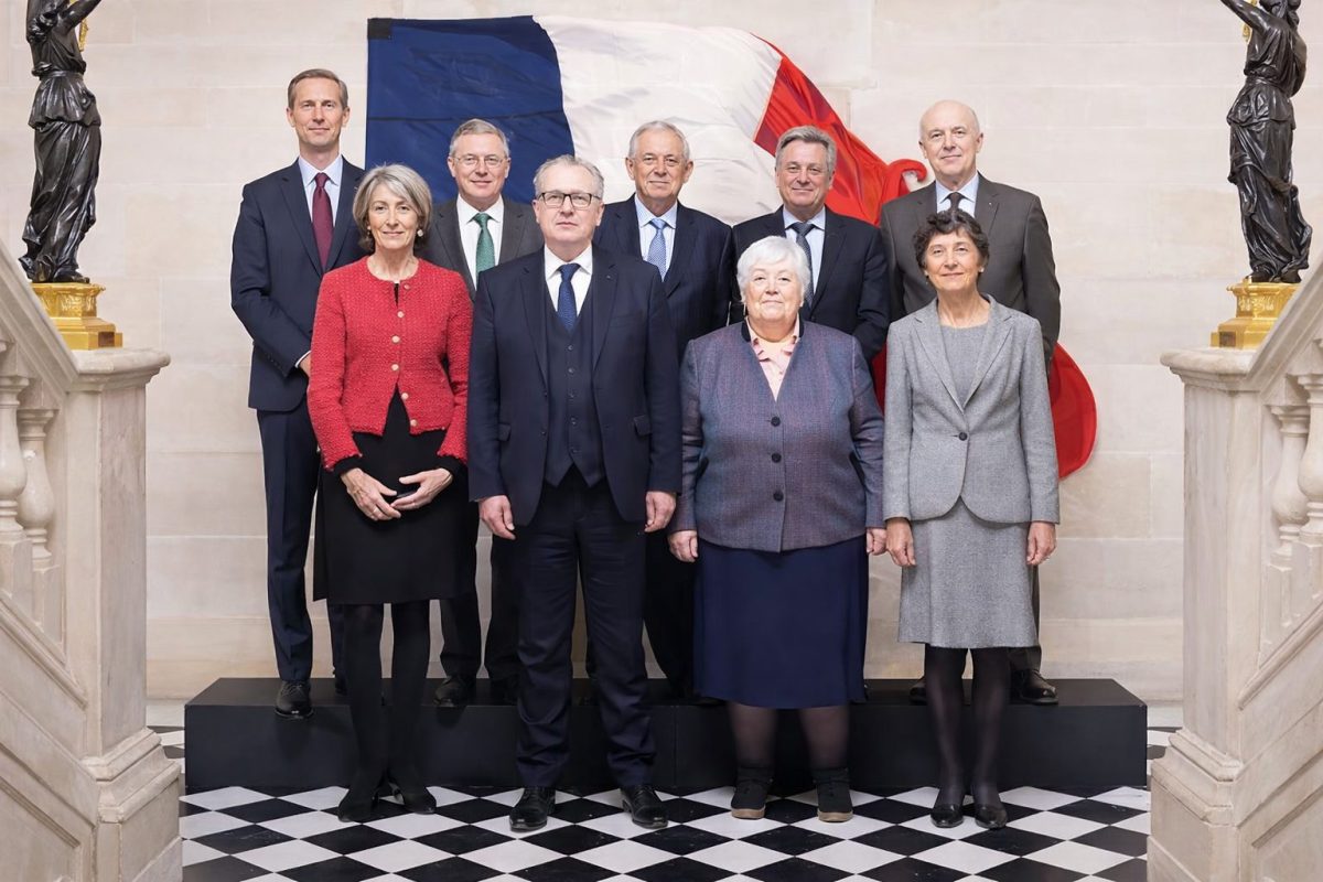 neuf personnes en tenue officielle posant en groupe dans un bâtiment institutionnel avec un drapeau tricolore en arrière-plan symbolisant une institution française importante