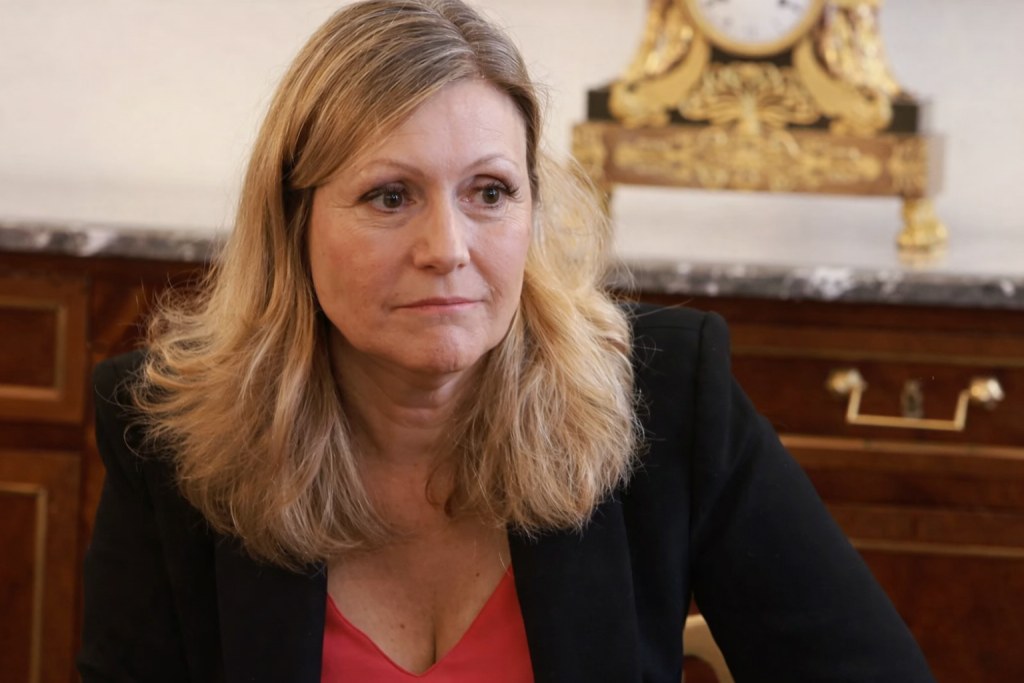 Portrait d’une femme politique assise dans un bureau officiel, regard sérieux, décor institutionnel en arrière-plan.