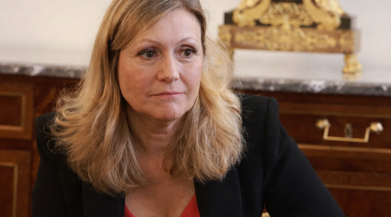 Portrait d’une femme politique assise dans un bureau officiel, regard sérieux, décor institutionnel en arrière-plan.